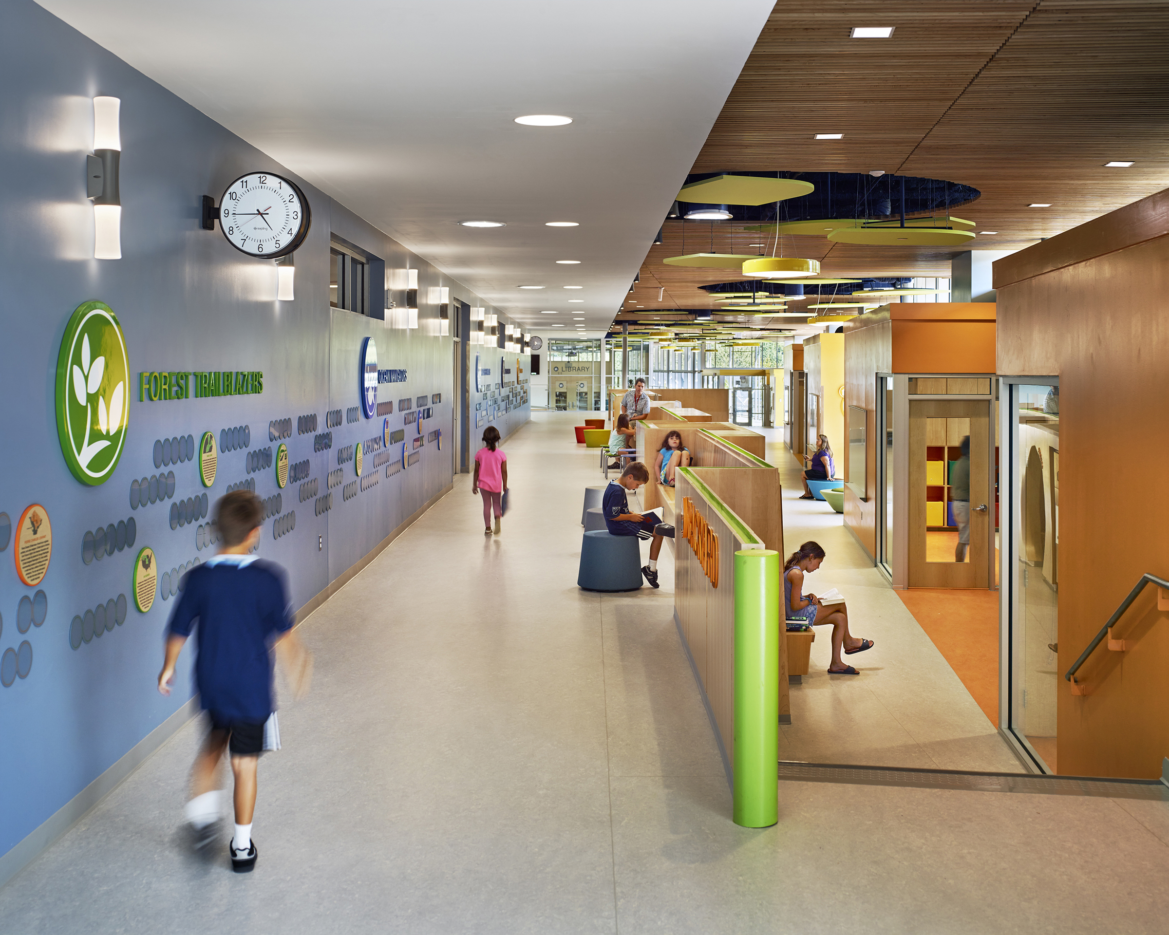 Two children walk down a hallway at Discovery Elementary School with acrylic wall graphics to their left while other children are sitting and reading to their right.