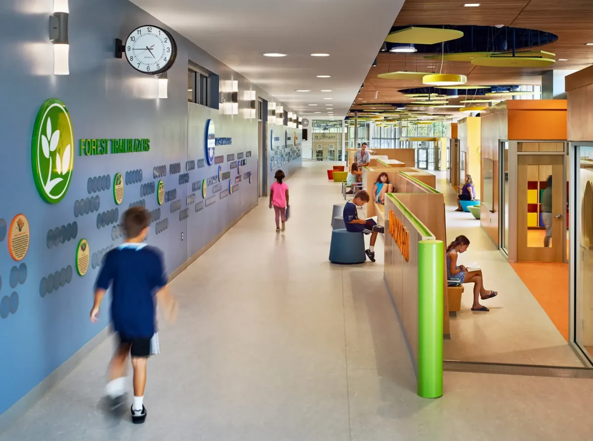 Two children walk down a hallway at Discovery Elementary School with acrylic wall graphics to their left while other children are sitting and reading to their right.