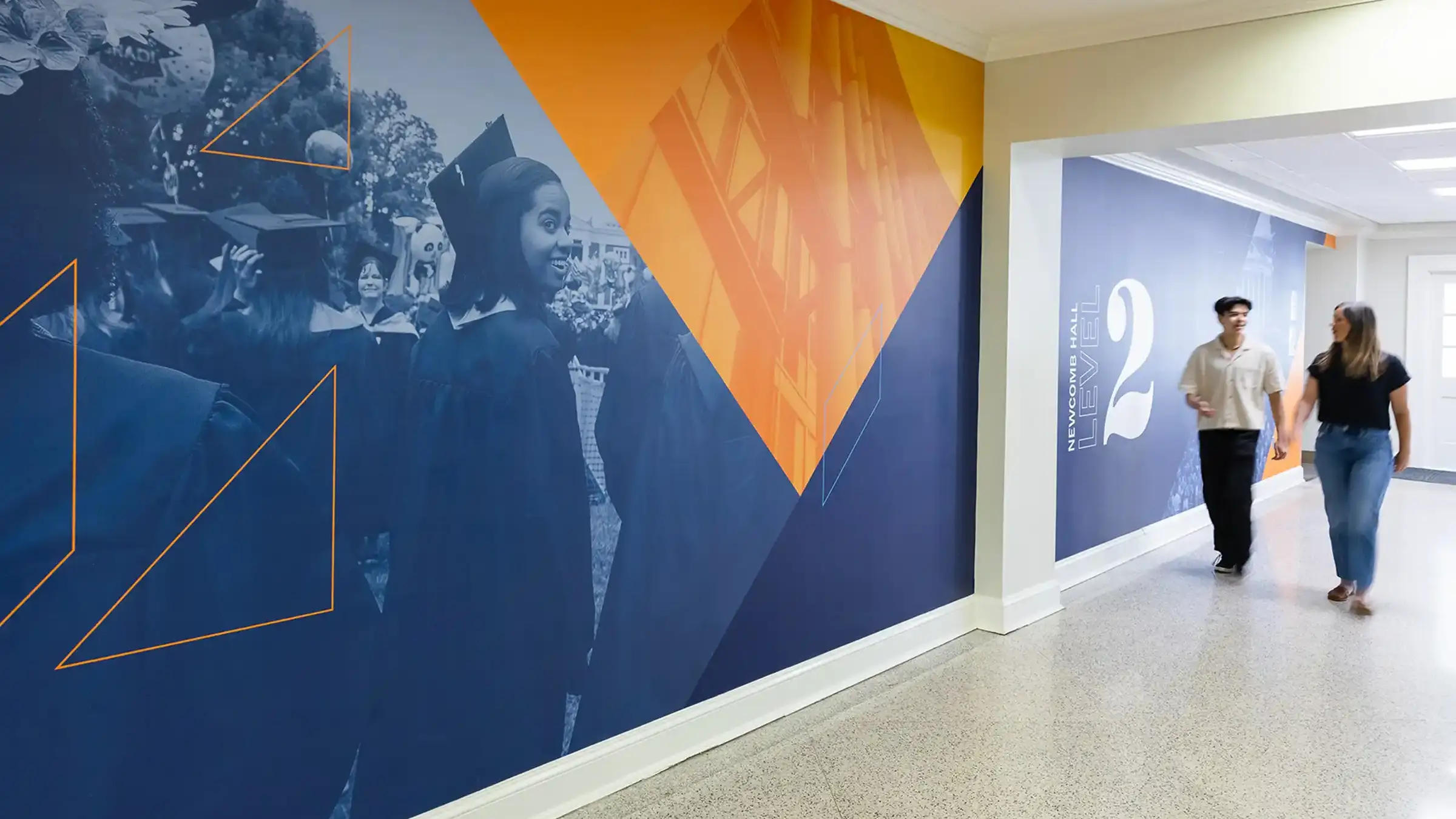 Two people walking down the hallway at Newcomb Hall with blue and orange wall graphics on the left side of the photo