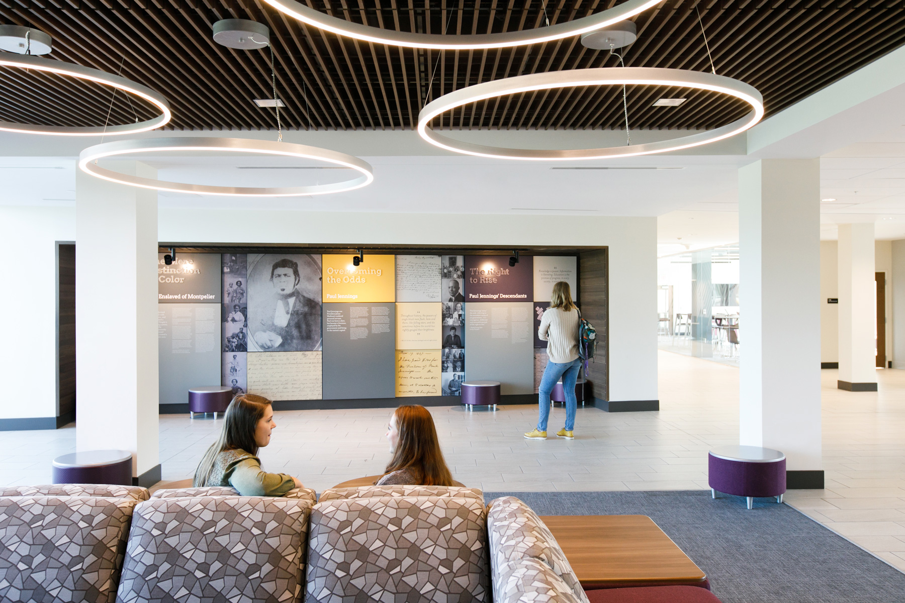 Two women walk on a couch while another woman reads exhibit displays about Paul Jennings at Jennings Residence Hall