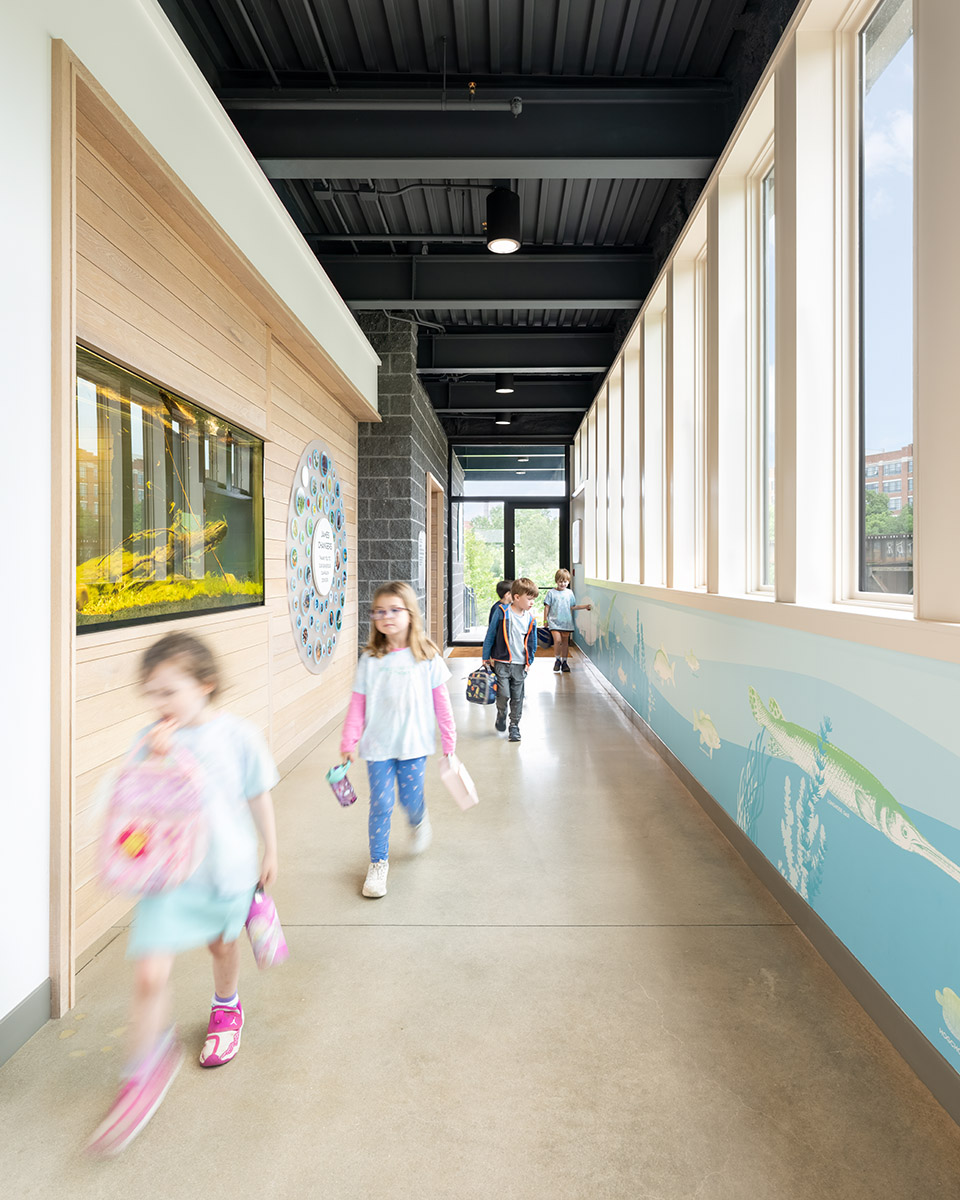 Children walk down the hallway at JRA Buzzard River Education Center past a donor wall on the left side of the photo and green and blue wall graphics on the right side.
