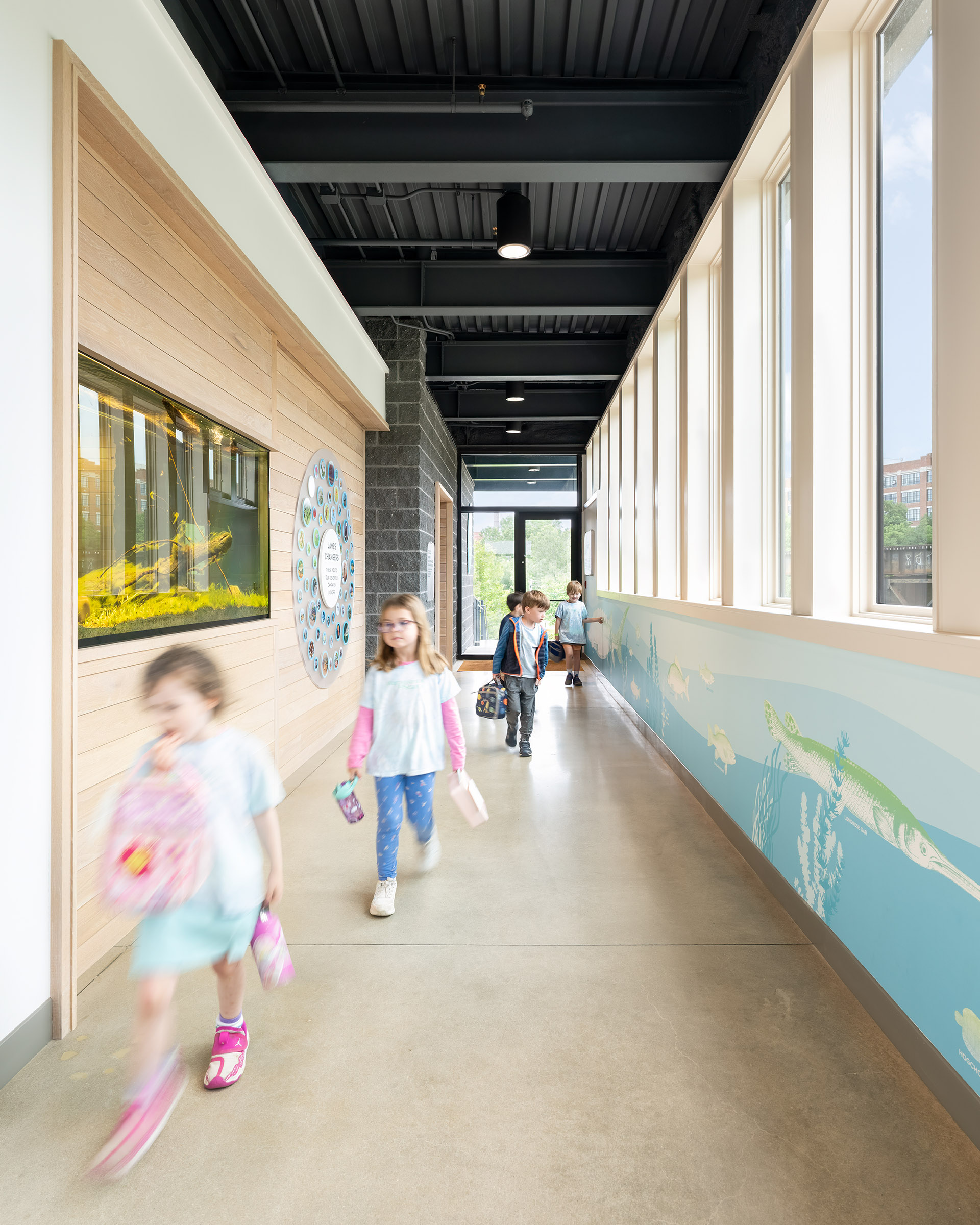 Children walk down the hallway at JRA Buzzard River Education Center past a donor wall on the left side of the photo and green and blue wall graphics on the right side.