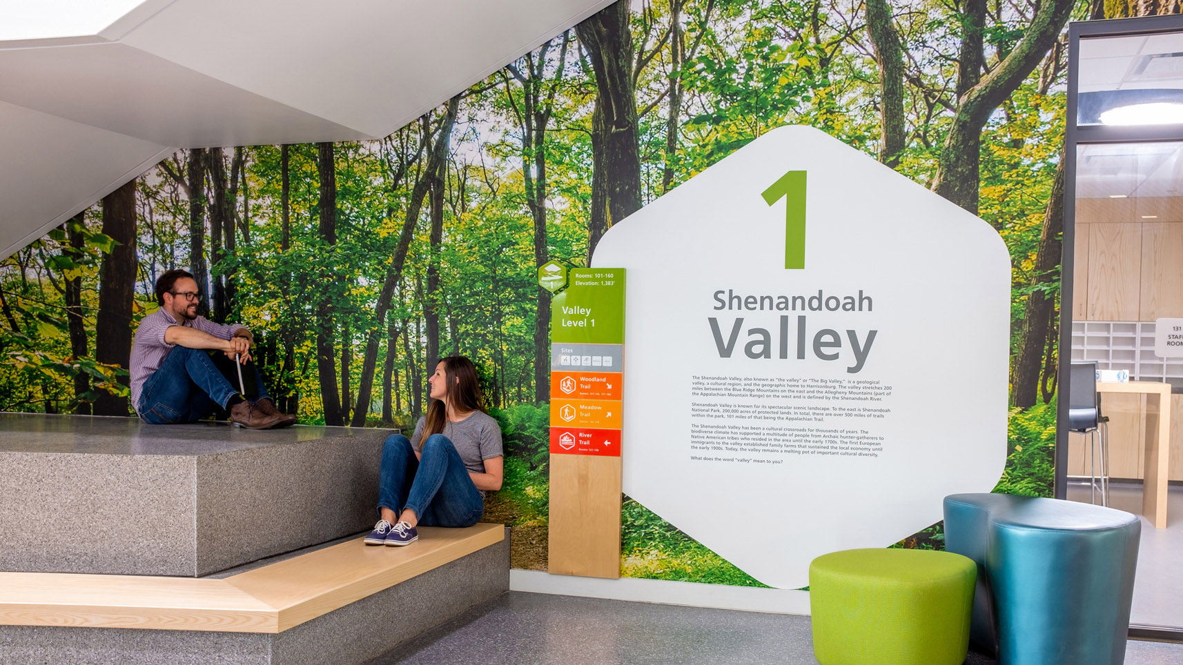 A man and a woman sit in front of wall graphics and wayfinding for floor level 1 at Bluestone Elementary School