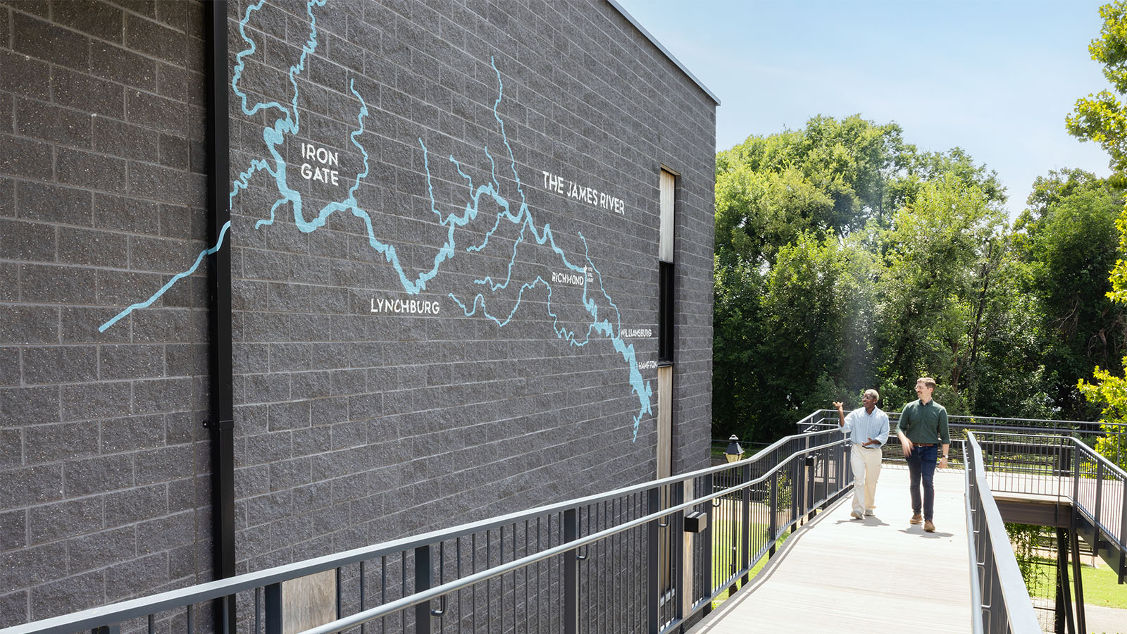 Two men walk up an outdoor ramp toward painted wall graphics of the James River at JRA Buzzard River Education Center